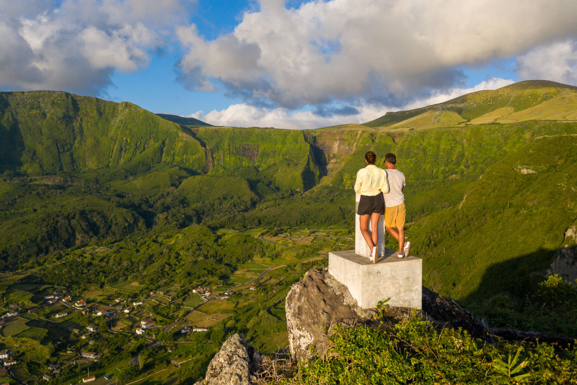 A couple stands at Miradouro do Portal above Faj&atilde;zinha on Flores Island in the Azores, Portugal. Steep green cliffs, narrow waterfalls, farmland and scattered houses fill the valley below.