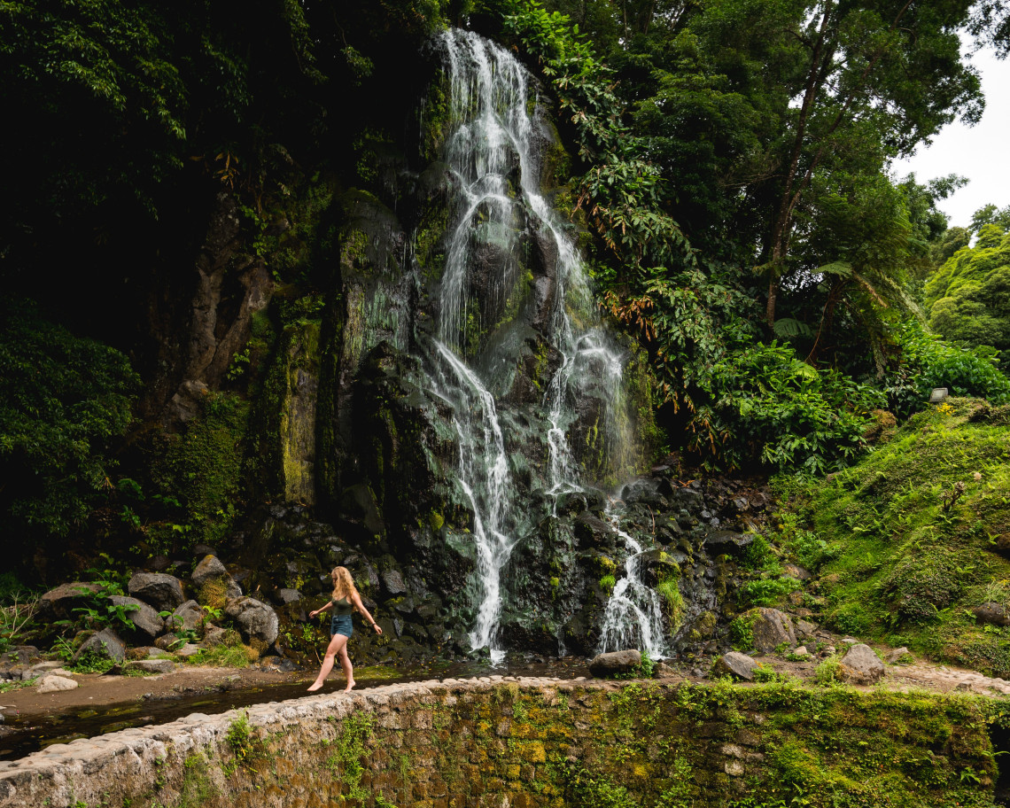 Waterfall cascading over dark volcanic rock at Ribeira dos Caldeir&otilde;es in Nordeste on S&atilde;o Miguel Island, surrounded by dense vegetation, with a visitor walking along the stone path below.