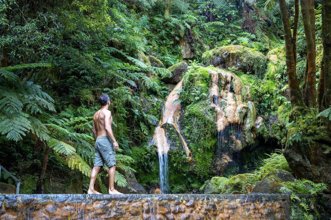 Visitor standing beside the warm waterfall at Caldeira Velha in Ribeira Grande, surrounded by giant ferns, moss-covered rock and dense forest.
