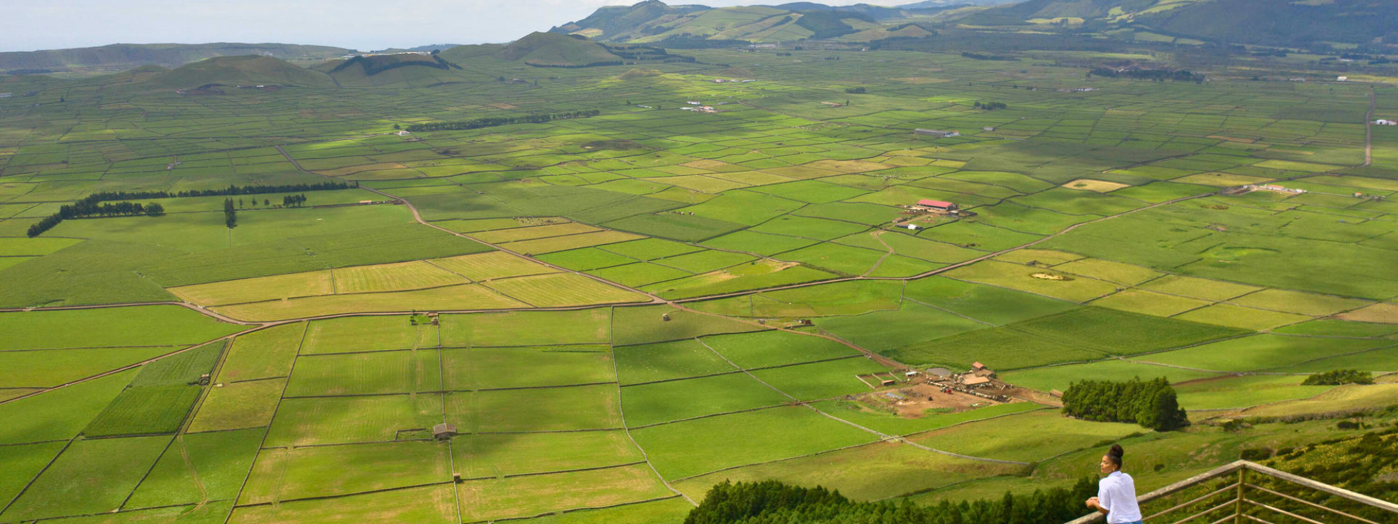 Solo visitor standing at the Serra do Cume viewpoint on Terceira Island, Azores, overlooking a vast patchwork of green fields divided by dark basalt stone walls, with rolling hills in the background