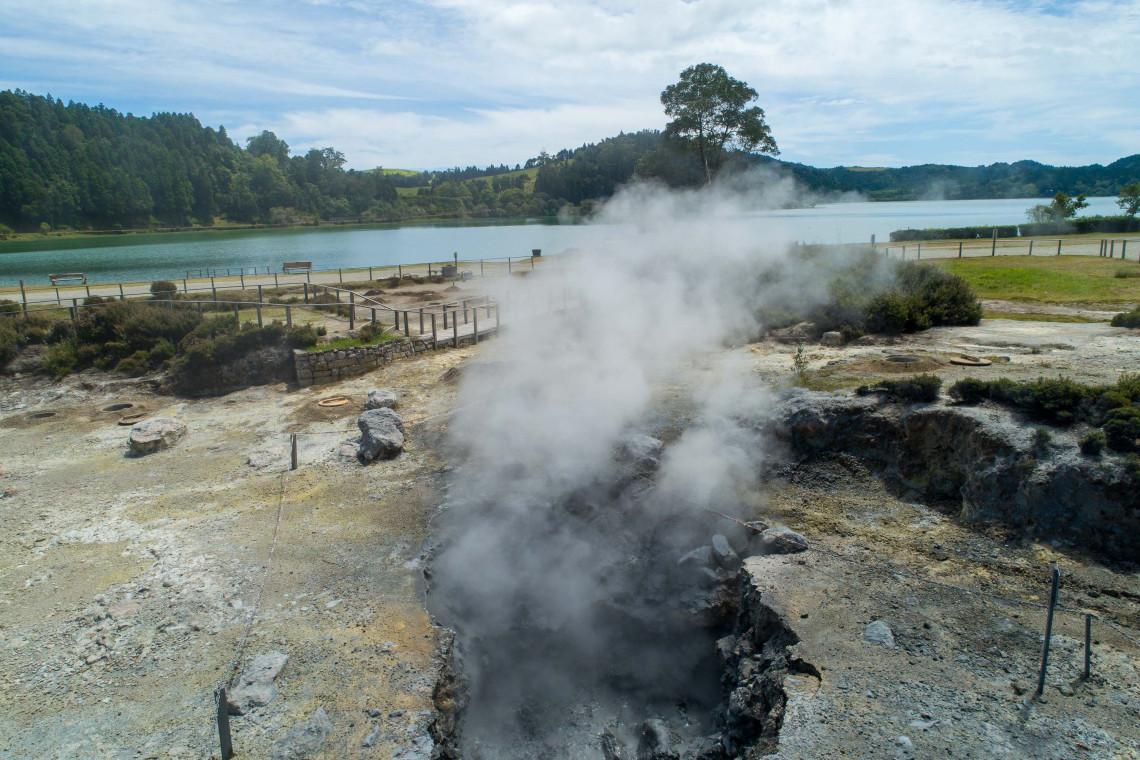 Steam rises from geothermal vents beside Lagoa das Furnas on S&atilde;o Miguel Island, surrounded by volcanic terrain, walking paths and green vegetation along the lakeshore.