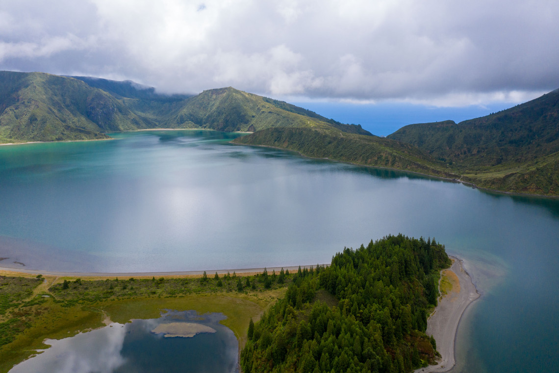 Aerial view of Lagoa do Fogo on S&atilde;o Miguel Island, showing the blue crater lake, narrow shoreline, forested slopes and steep volcanic ridges beneath low clouds.