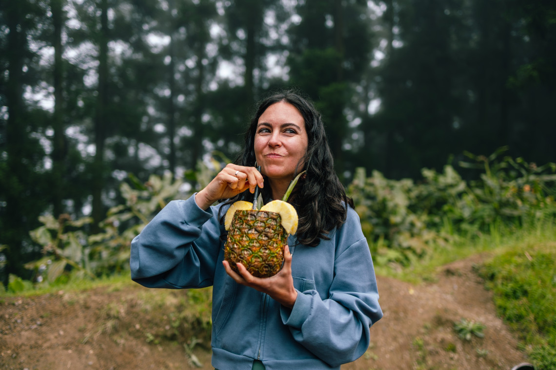 Visitor enjoying a fresh drink served inside a pineapple while exploring the green countryside of S&atilde;o Miguel Island in the Azores.