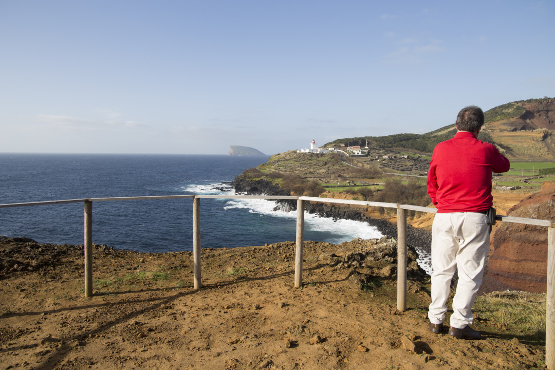 Man standing at a coastal viewpoint in the Azores overlooking cliffs, ocean, and a lighthouse, enjoying a peaceful solo travel experience