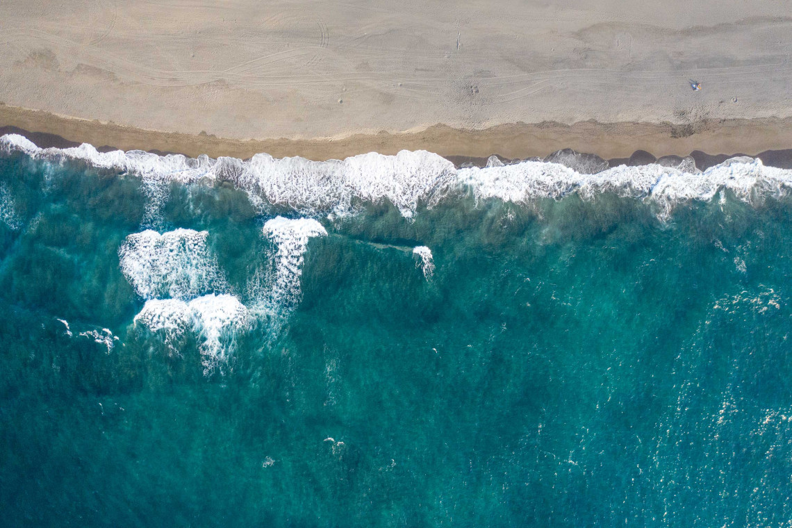 Overhead aerial view of Atlantic waves reaching a dark sandy beach in the Azores, with clear turquoise water revealing the shallow coastal seabed.