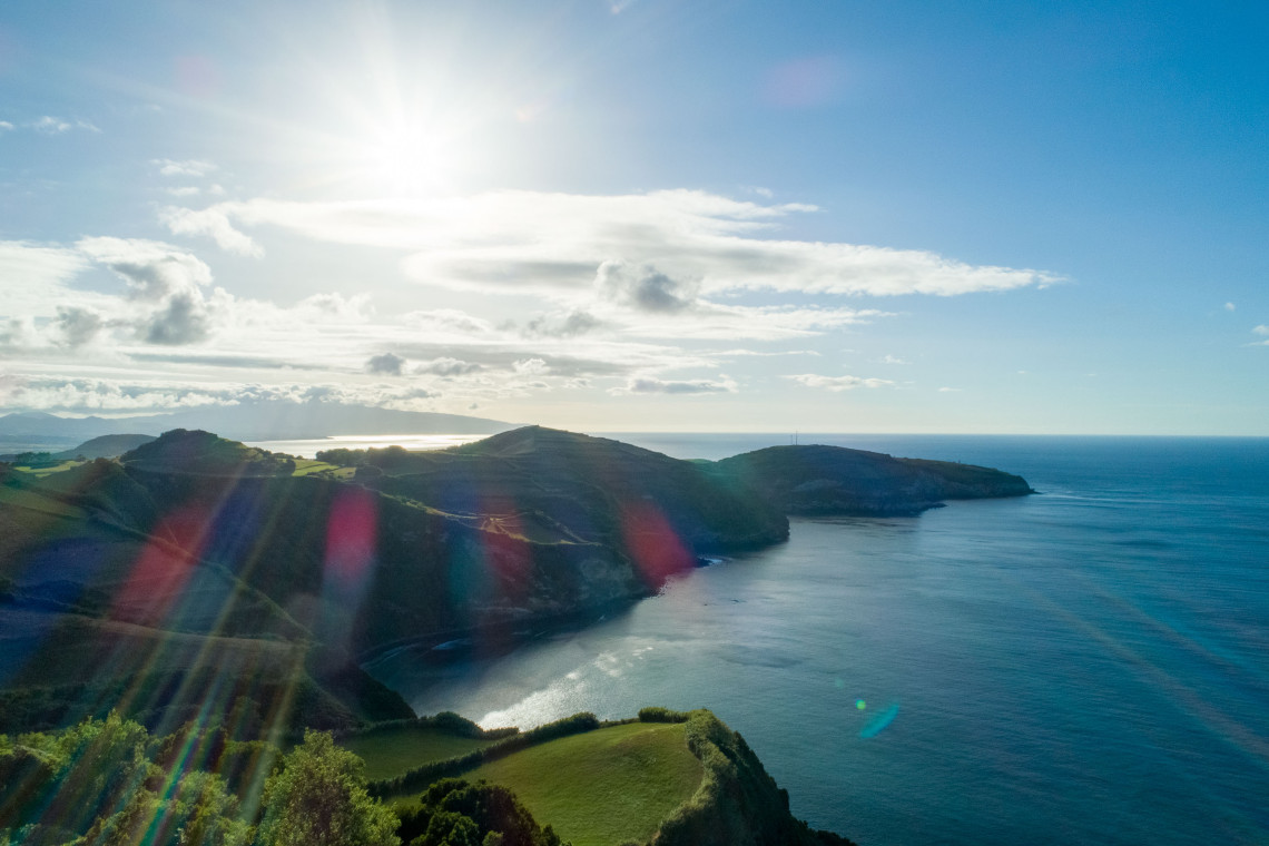 Sunlight shines over a quiet Atlantic bay bordered by green volcanic cliffs and rolling coastal hills in the Azores.