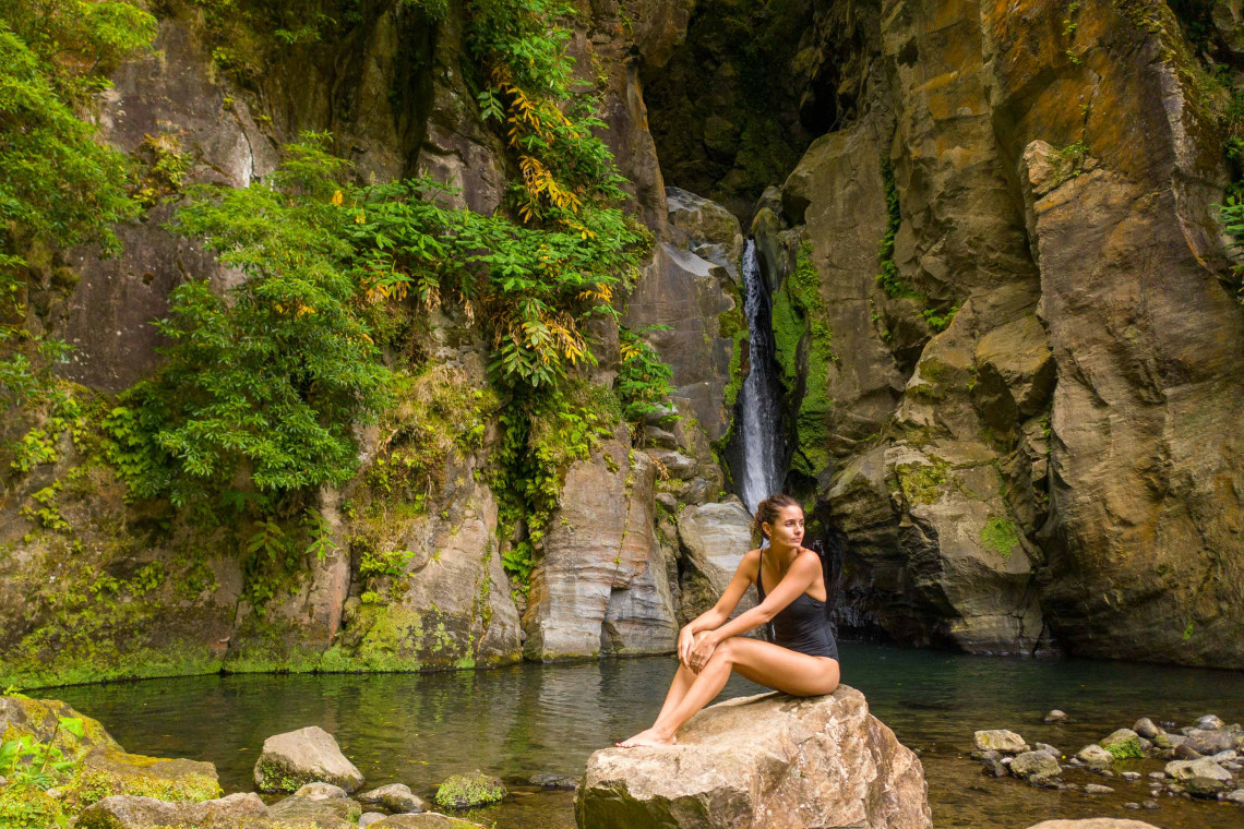 A visitor sits beside the natural pool below Salto do Cabrito waterfall near Ribeira Grande, surrounded by steep volcanic rock walls and dense vegetation.