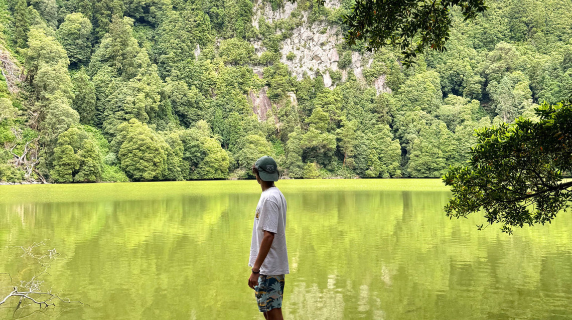 A visitor stands beside Lagoa do Congro on S&atilde;o Miguel Island, where green crater waters are surrounded by steep slopes covered in dense native and subtropical forest.