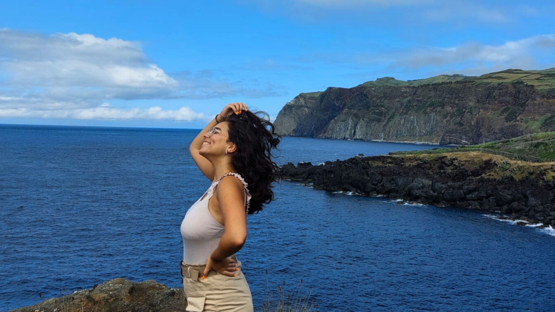A visitor enjoys a viewpoint along the coast of Graciosa Island, overlooking deep blue Atlantic waters, dark volcanic rocks and steep green coastal cliffs.