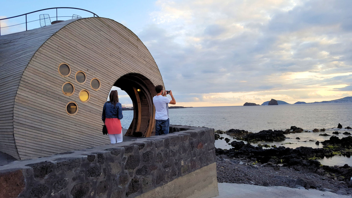 Visitors explore Cella Bar on the waterfront of Madalena on Pico Island, with its distinctive curved wooden architecture overlooking the Atlantic Ocean and neighboring islands.