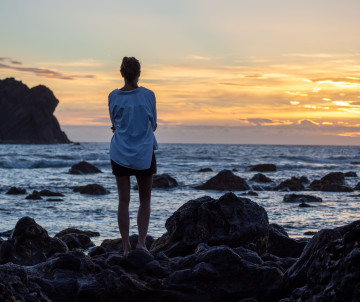 A visitor stands on the dark volcanic shoreline of Mosteiros on São Miguel Island, watching the sun set over the Atlantic Ocean and offshore rock formations.