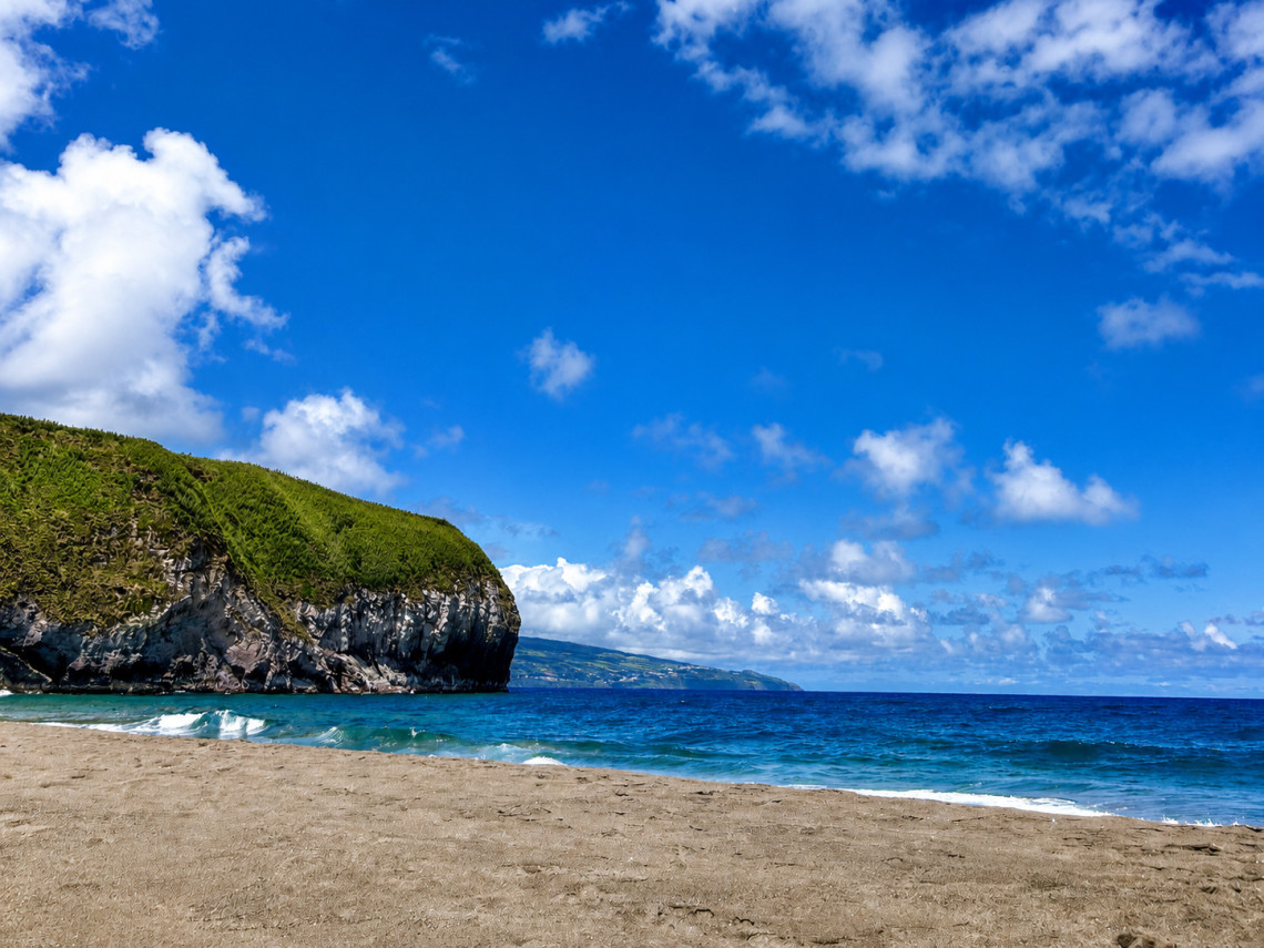  Sandy beach at Areal de Santa B&aacute;rbara on S&atilde;o Miguel Island, Azores, with a dramatic green-topped basalt headland on the left, turquoise Atlantic water, and a neighbouring island visible on the horizon
