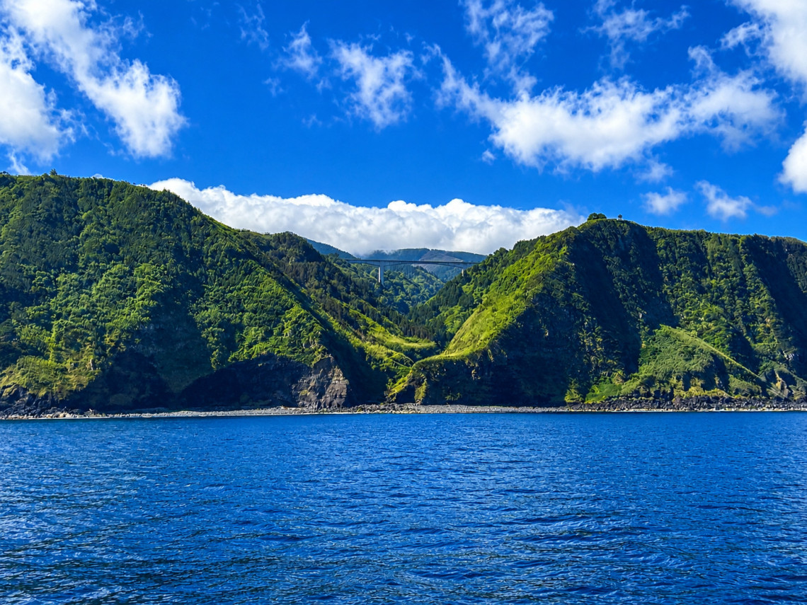 View from the ocean of the dramatic green volcanic sea cliffs of Nordeste, S&atilde;o Miguel Island, Azores, with a bridge visible between two lush ridges and dark basalt rock at the waterline