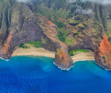 Aerial view of the rugged Nā Pali Coast on Kauai, Hawaii, showing steep red volcanic cliffs and narrow sandy beach meeting deep blue ocean water