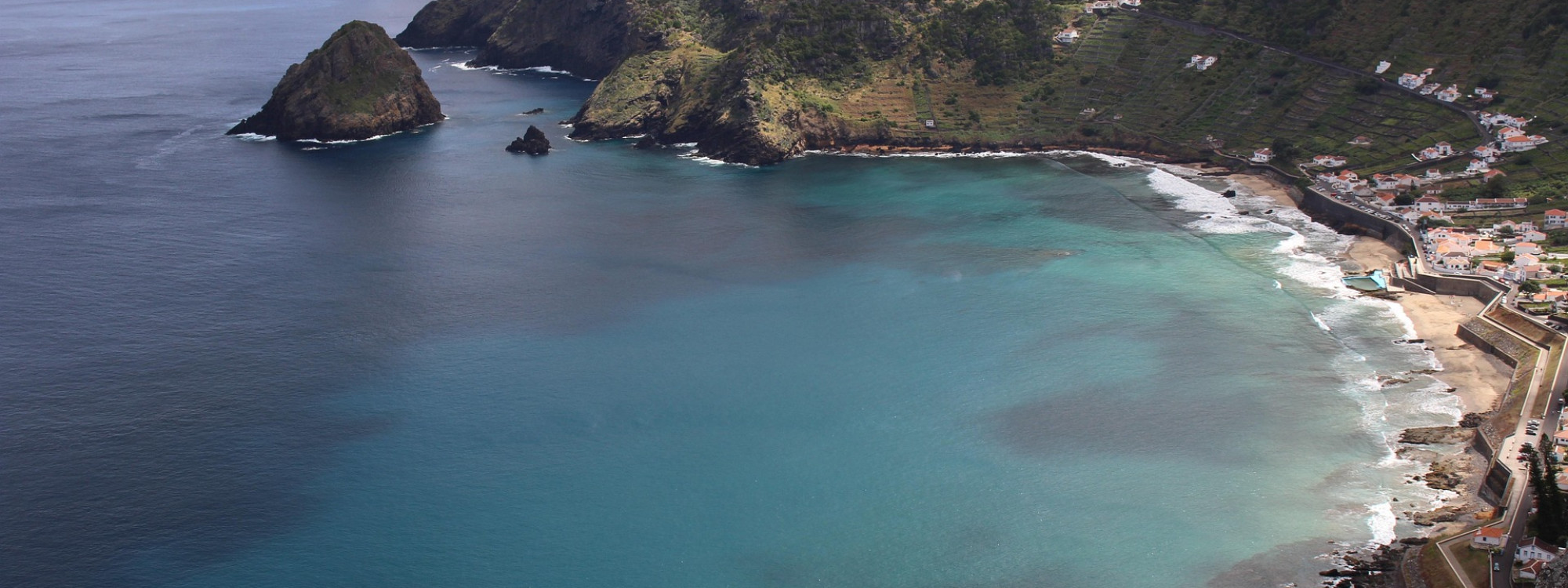 Aerial view of São Lourenço Bay on Santa Maria Island, showing clear blue Atlantic waters, steep green slopes, terraced vineyards and the coastal settlement lining the shore.