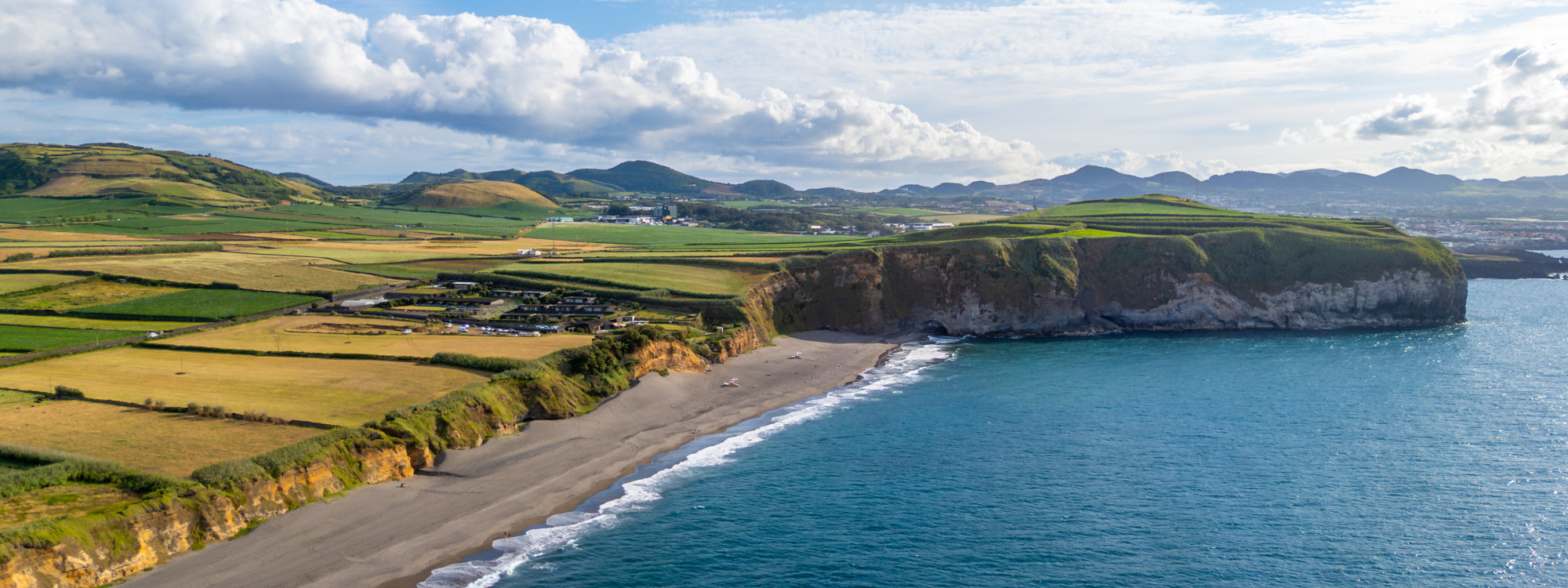 Wide aerial view of Praia de Santa Bárbara dark sand beach with Atlantic waves breaking on the shore, dramatic green cliffs, and clear blue sky, north coast of São Miguel Island, Azores
