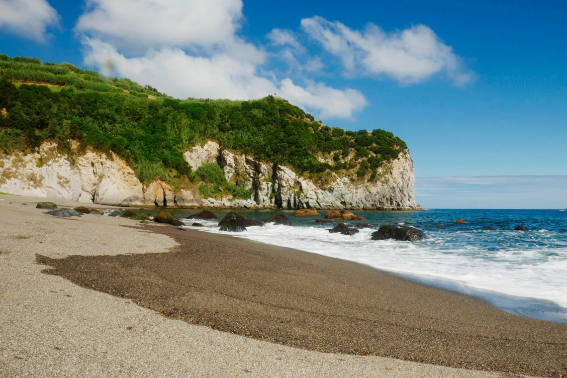 Dark sand beach at Praia dos Moinhos in Porto Formoso with Atlantic waves, dramatic cliffs covered in green vegetation, and clear blue sky, north coast of S&atilde;o Miguel Island, Azores