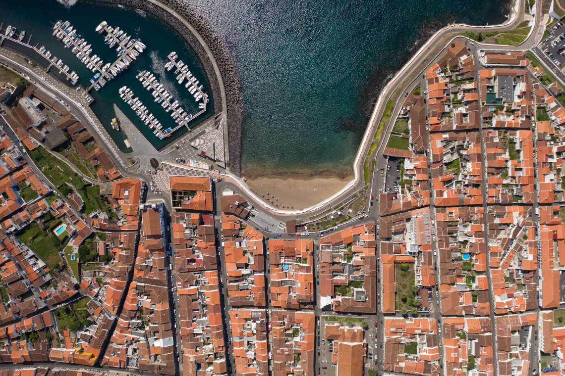 Aerial top-down view of Prainha city beach tucked into a sheltered bay in Angra do Hero&iacute;smo, with the marina, waterfront promenade, and terracotta rooftops of the UNESCO city, Terceira Island, Azores