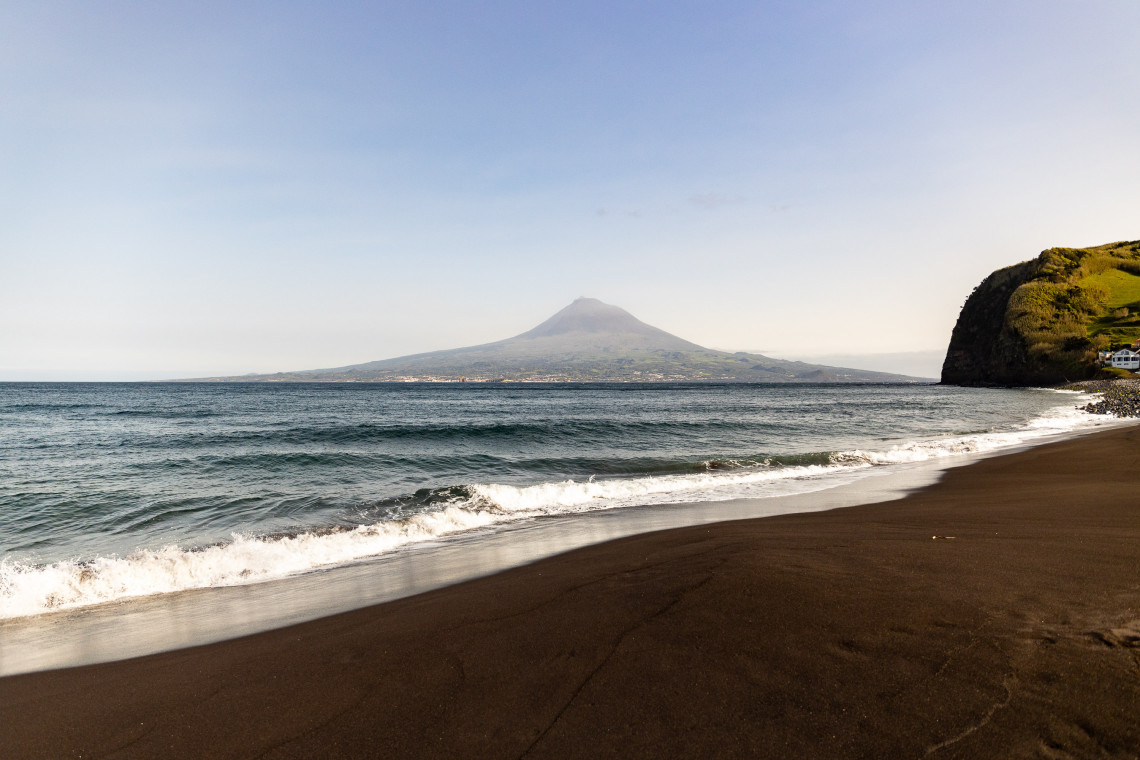 Dark sand beach at Praia de Almoxarife on the north coast of Faial Island, with the iconic cone of Mount Pico rising dramatically across the channel in the background, Azores