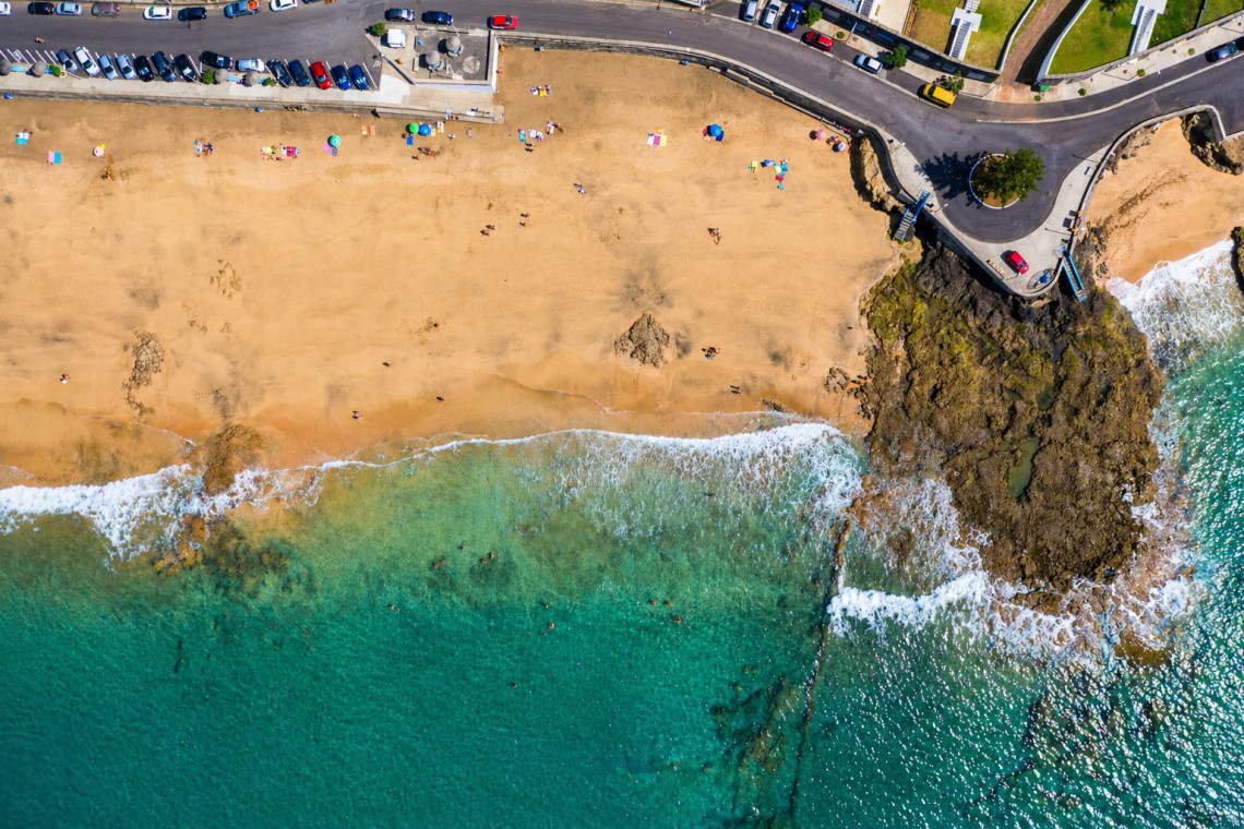 Aerial top-down view of Praia Formosa golden sand beach with turquoise Atlantic water, sunbathers, and coastal road, Santa Maria Island, Azores
