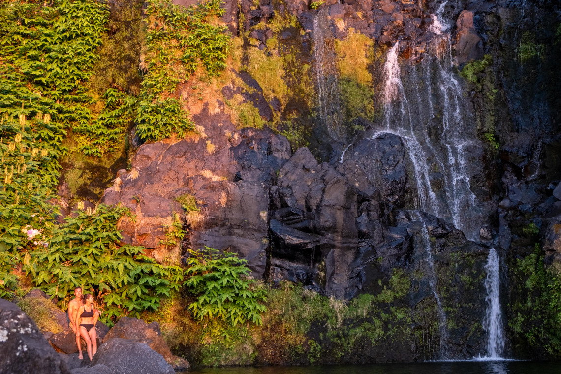Couple sitting beside the dramatic Cascata do Po&ccedil;o do Bacalhau waterfall cascading down a lush green volcanic cliff face into a natural pool, Flores Island, Azores