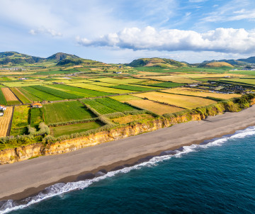 Aerial view of Praia de Santa Bárbara dark sand beach with Atlantic waves, patchwork green fields, and volcanic hills in the background, north coast of São Miguel Island, Azores