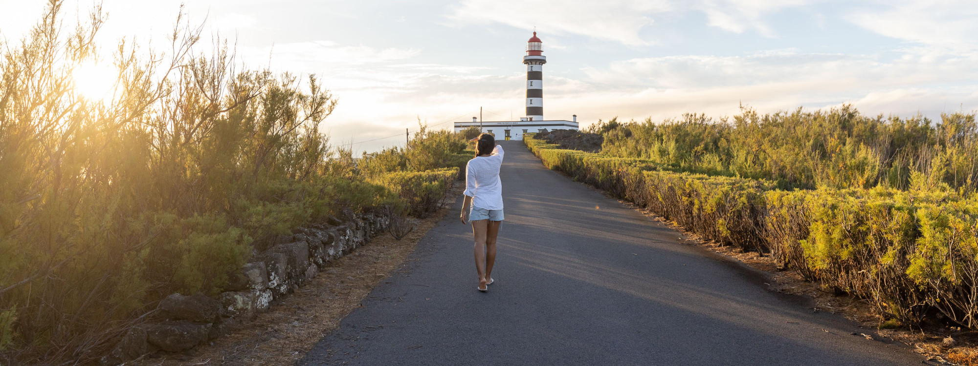 Traveler walking along the road toward the black-and-white Ponta da Barca Lighthouse on Graciosa Island, surrounded by low coastal vegetation in the evening light.
