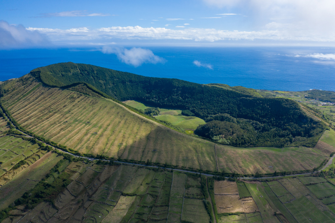 Aerial view of Caldeira da Graciosa, showing the broad volcanic crater covered by forest and surrounded by agricultural fields with the Atlantic Ocean beyond.