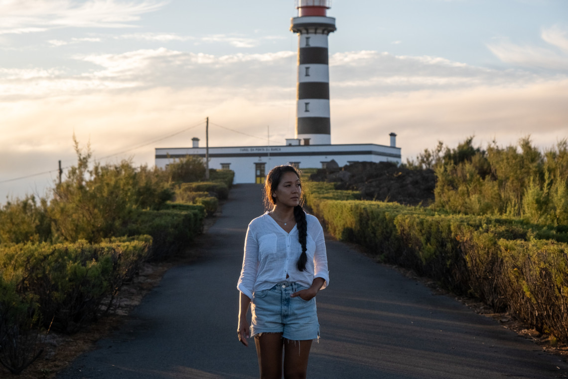 Traveler walking in front of Ponta da Barca Lighthouse on Graciosa Island, with the striped lighthouse tower rising above coastal vegetation in warm evening light.