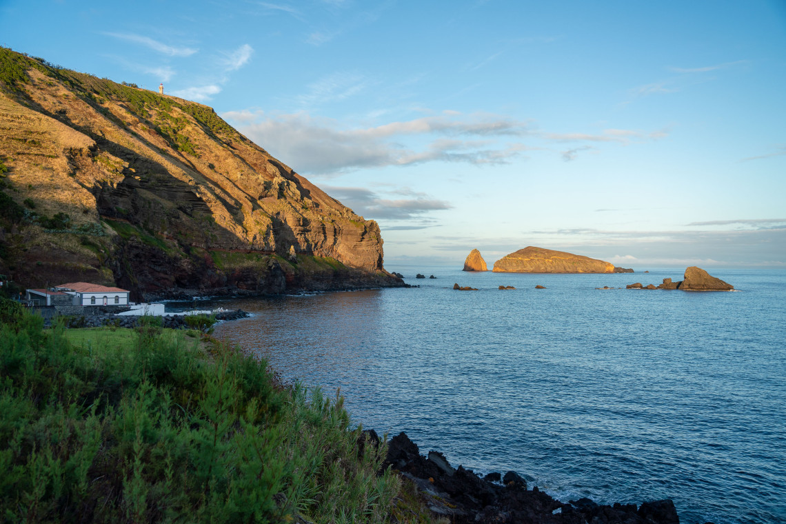 View across the Atlantic toward the rocky Ilh&eacute;us do Carapacho off the southern coast of Graciosa Island, framed by layered volcanic cliffs and coastal vegetation. The islets lie near the Carapacho bathing area and Ponta da Restinga.
