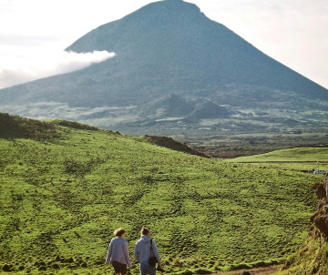 Mount Pico rises above green volcanic terrain on Pico Island as two hikers walk through the landscape below Portugal’s highest mountain.