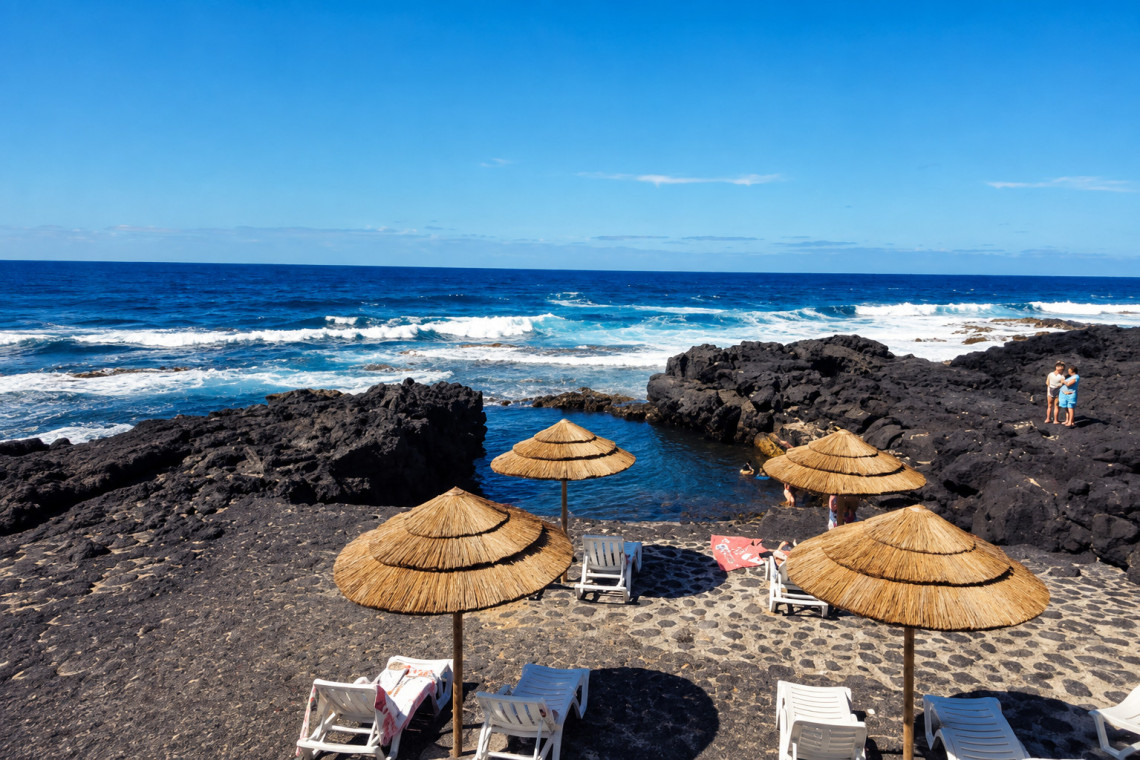 Cais do Mourato natural swimming pool on Pico Island, with black volcanic rock, straw sun umbrellas and direct views of the Atlantic Ocean near Bandeiras, Azores.