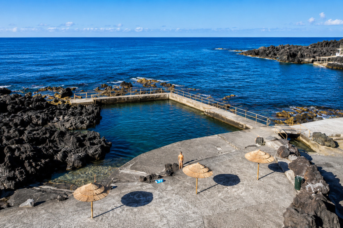 Piscina de S&atilde;o Mateus on Pico Island, a natural ocean swimming area bordered by black volcanic rock and concrete platforms overlooking the Atlantic. Straw sun umbrellas and direct sea access surround the protected pool.