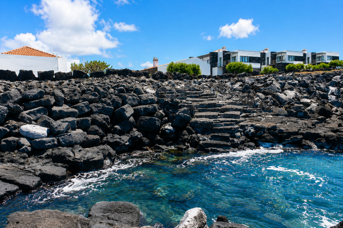 Stone steps descend through black volcanic rock toward the Atlantic at Barca in Madalena, Pico Island. Traditional and modern buildings line the coastal settlement above the water.
