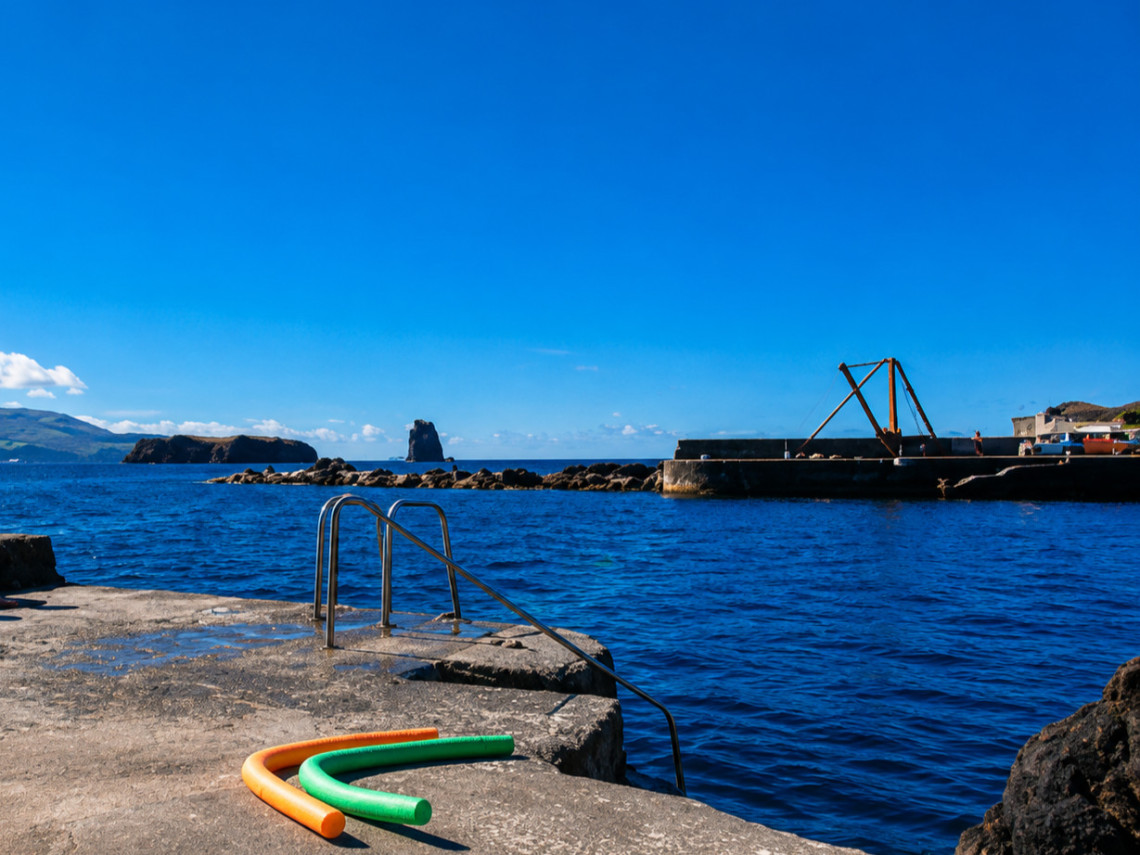 Porto da Areia Larga bathing area in Madalena on Pico Island, with a metal swimming ladder leading directly into the Atlantic beside the harbor and volcanic coastline.