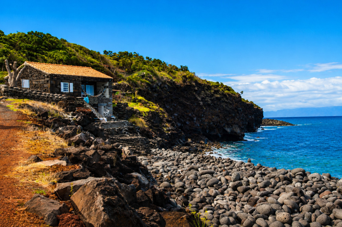 Ba&iacute;a das Canas in Prainha de Cima on Pico Island, with a shoreline of rounded volcanic stones below a traditional stone house and green coastal slopes overlooking the Atlantic.