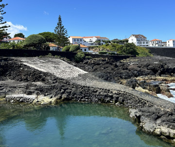Natural seawater pool surrounded by dark volcanic rock on Pico Island in the Azores, Portugal, with traditional houses and coastal vegetation in the background.