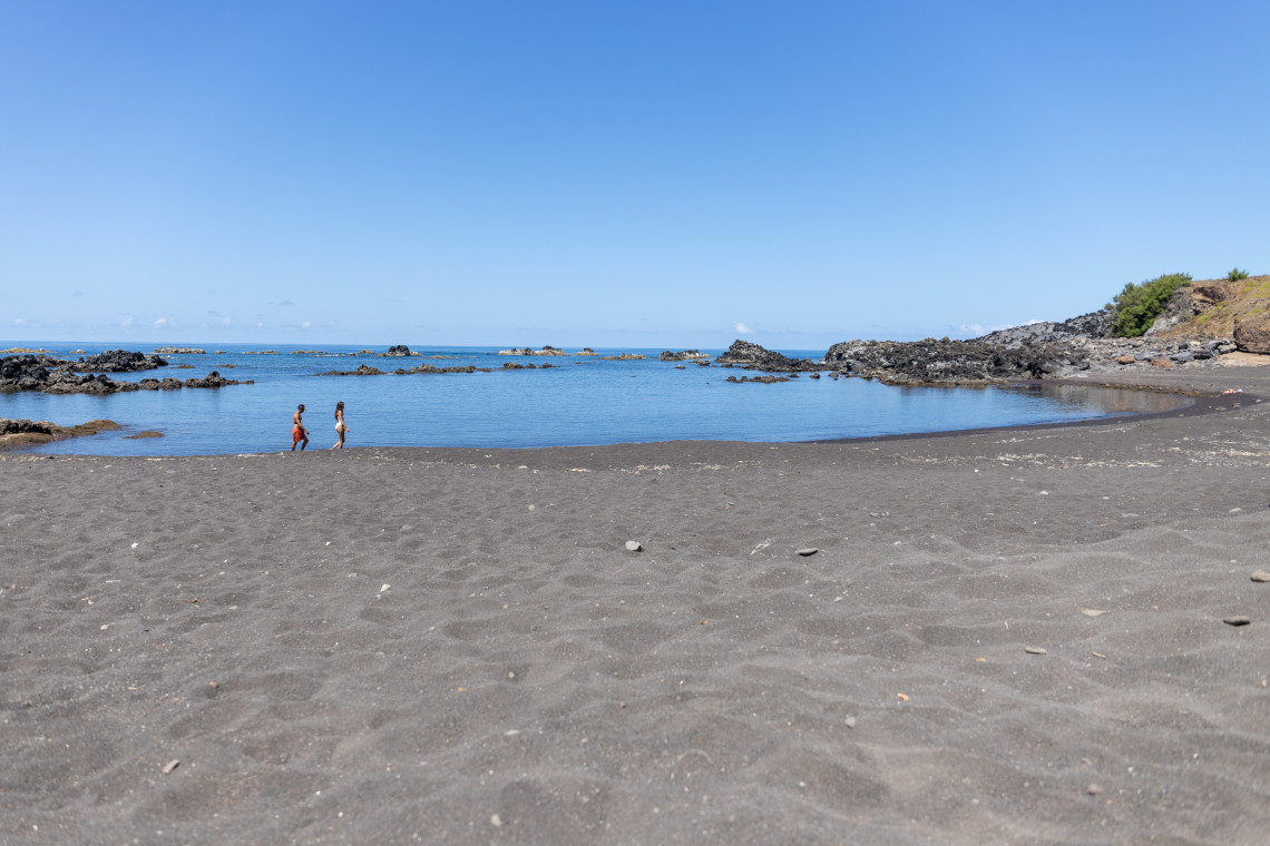 Praia da Areia in Vila do Corvo features dark volcanic sand and calm Atlantic water surrounded by basalt rock formations. Two visitors walk along the shoreline of Corvo Island in the Azores, Portugal.
