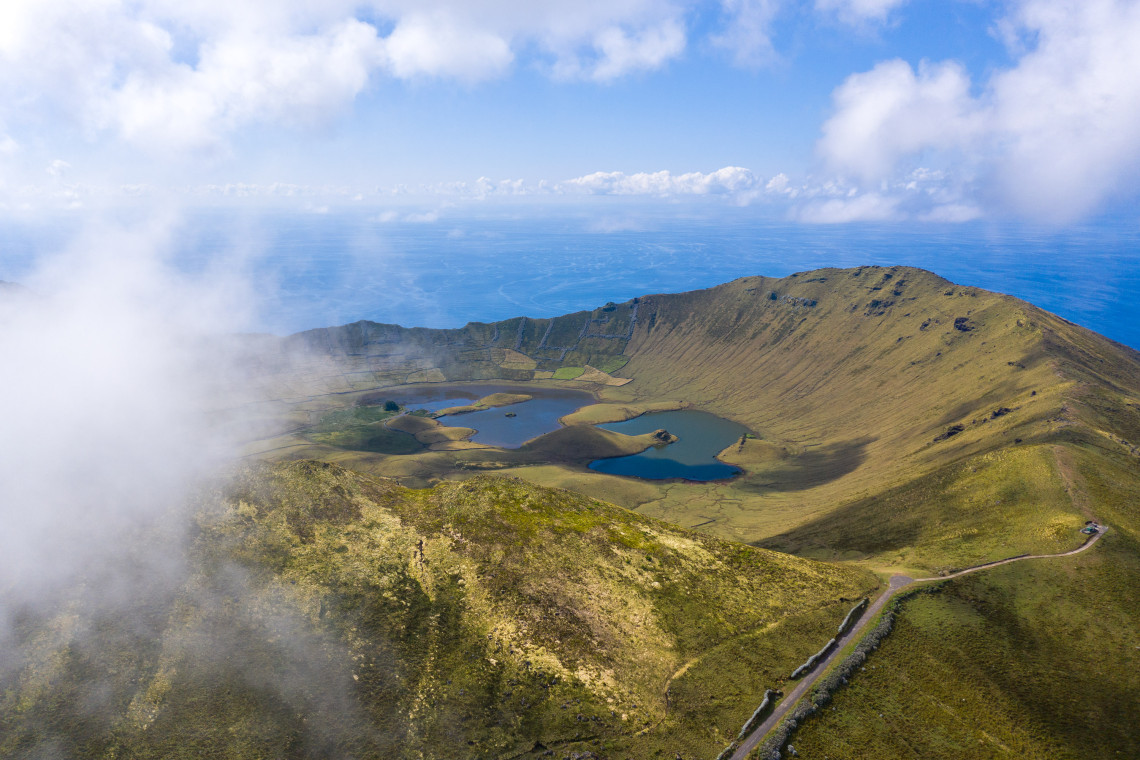 Aerial view of Caldeir&atilde;o Crater on Corvo Island in the Azores, Portugal, with clouds drifting across the caldera rim. Small lakes and volcanic formations spread across the green crater floor, with the Atlantic Ocean beyond.