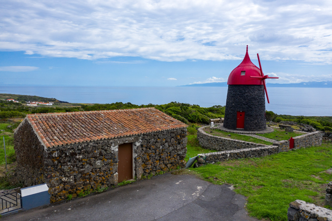 Traditional basalt windmill on Graciosa Island with a distinctive red wooden cap, standing beside a stone cottage and low volcanic walls overlooking the Atlantic.
