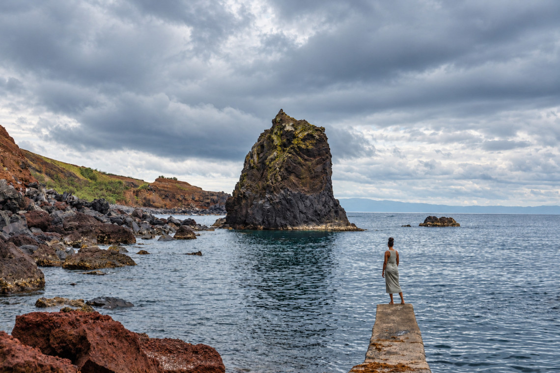 Traveler overlooking Porto Afonso on Graciosa Island, where a tall volcanic sea stack rises from a sheltered cove surrounded by layered red and dark rock cliffs.