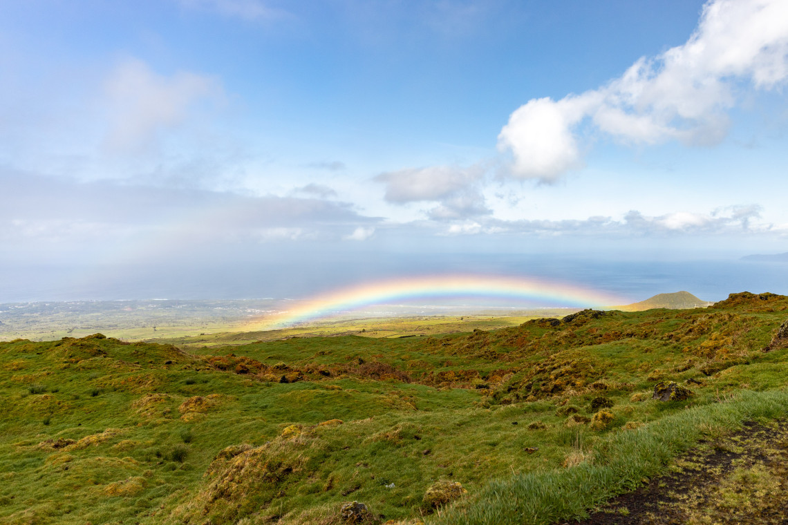 Rainbow stretching across the green volcanic highlands of Pico Island beneath low clouds, with the Atlantic Ocean visible beyond the slopes.