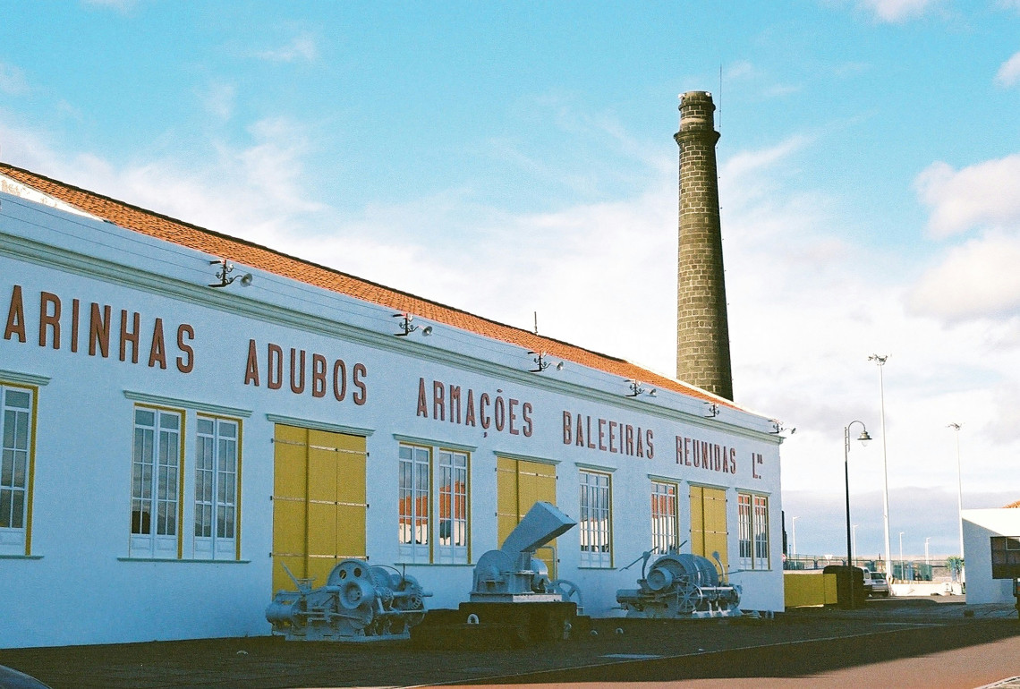 Exterior of the Museu da Ind&uacute;stria Baleeira in S&atilde;o Roque do Pico, housed in the former Arma&ccedil;&otilde;es Baleeiras Reunidas whaling factory, with industrial machinery and its tall chimney visible outside.