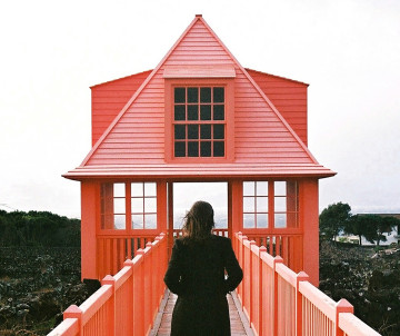 Visitor walking along the raised wooden walkway toward the red mirante at the Museu do Vinho in Madalena, surrounded by Pico Island’s volcanic vineyard landscape.
