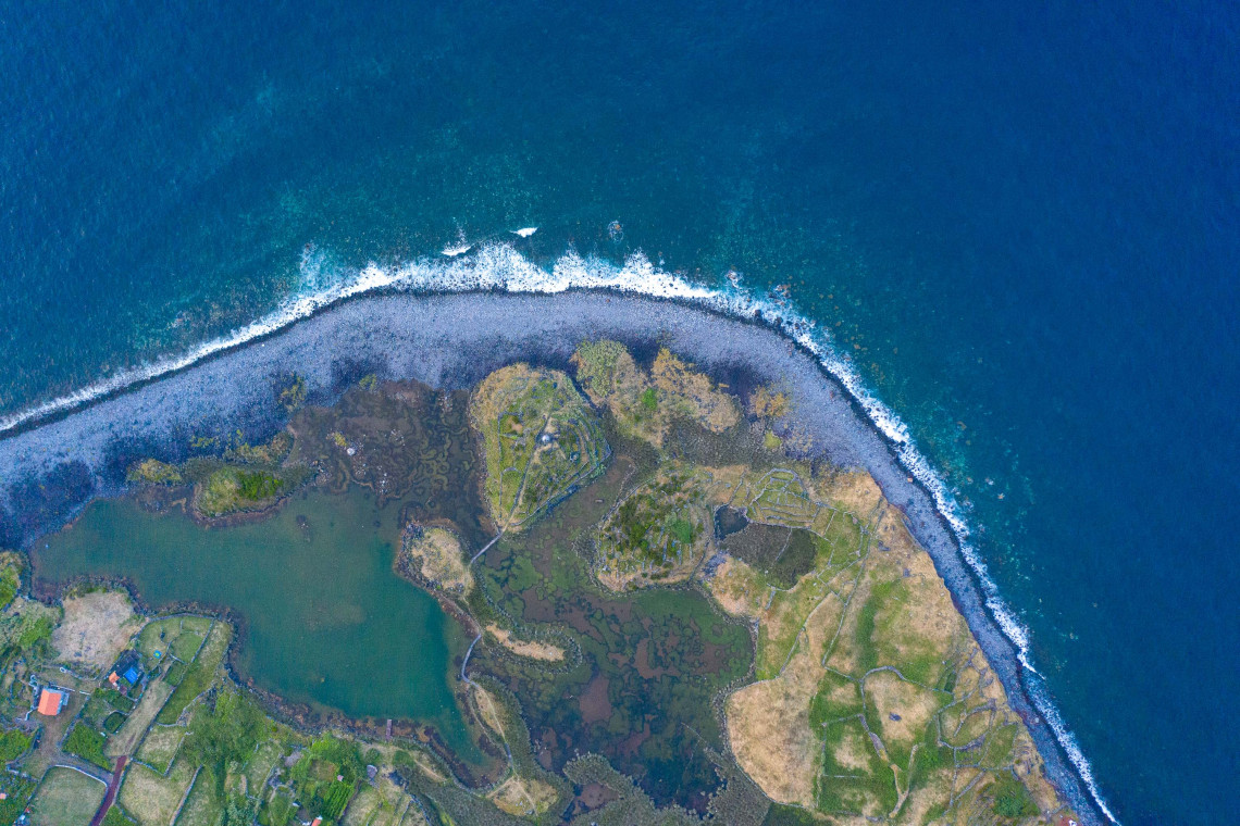 Top-down aerial view of Faj&atilde; dos Cubres on S&atilde;o Jorge Island, showing its coastal lagoon, wetlands and dark volcanic shoreline beside the Atlantic Ocean.
