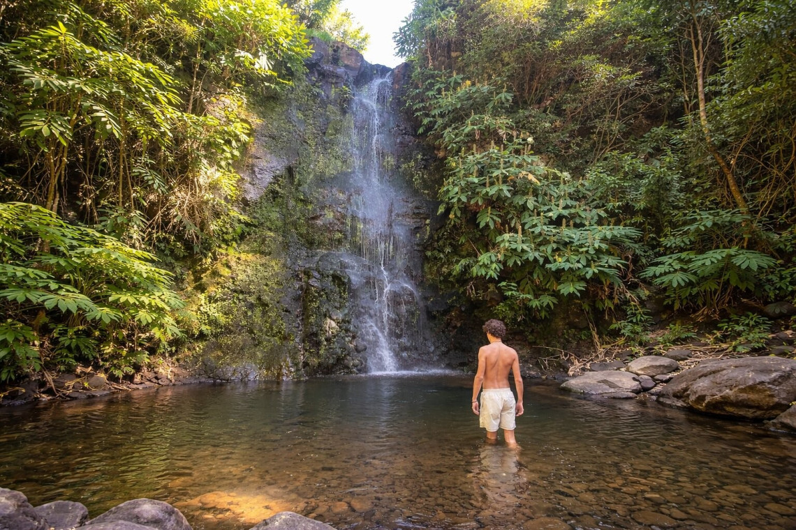 Traveler standing in the natural pool below Cascata do Cruzal on S&atilde;o Jorge Island, surrounded by dense vegetation and volcanic rock.