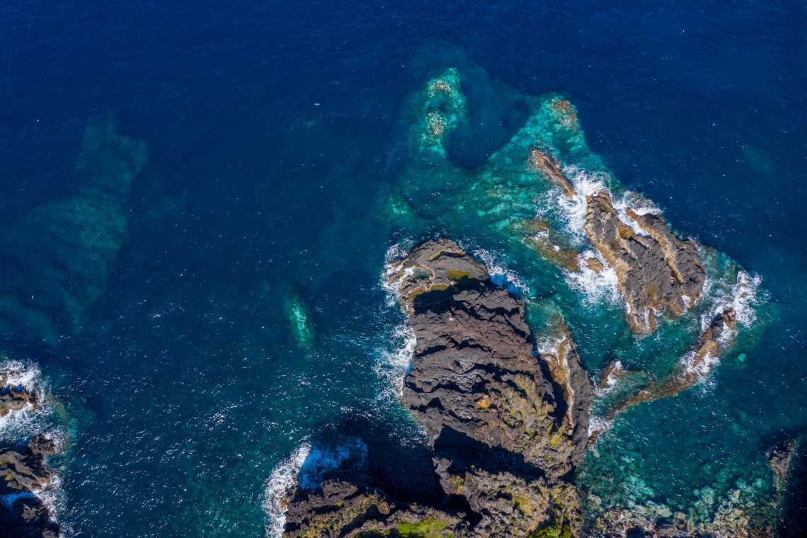 Aerial view of jagged basalt rock formations surrounded by clear blue Atlantic water along the volcanic coast of S&atilde;o Jorge Island.