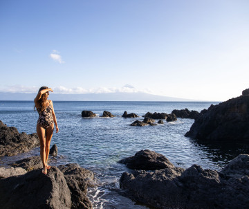 Traveler standing on dark volcanic rocks along the coast of São Jorge Island, with the Atlantic Ocean and Mount Pico visible in the distance.