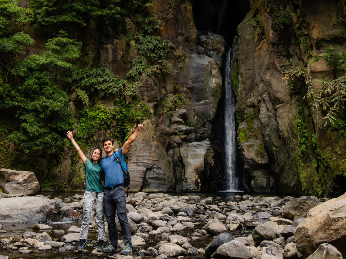 Couple standing beneath Salto do Cabrito waterfall near Ribeira Grande, surrounded by steep volcanic rock walls and lush vegetation.