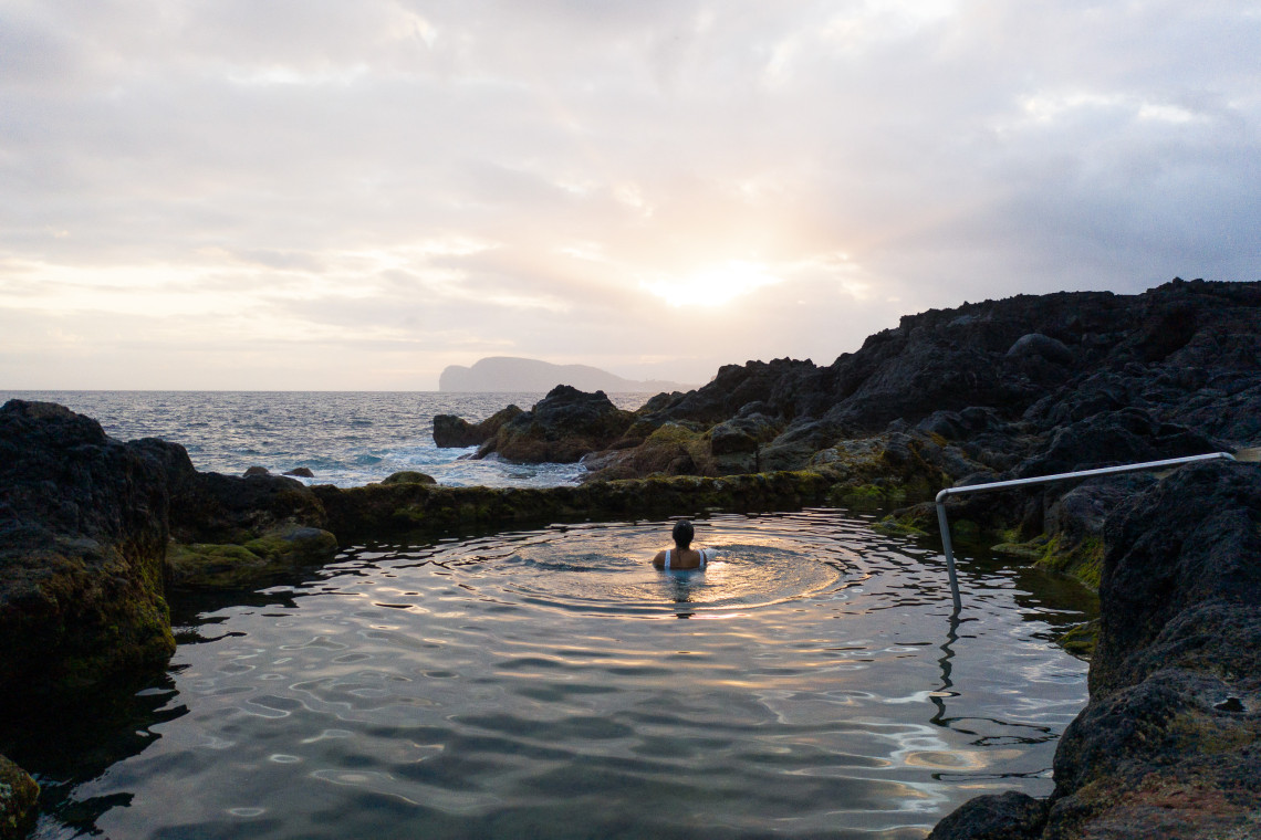Person swimming in the natural volcanic pools of Serretinha on Terceira Island, Azores, surrounded by dark lava rock as the sun sets over the Atlantic Ocean.